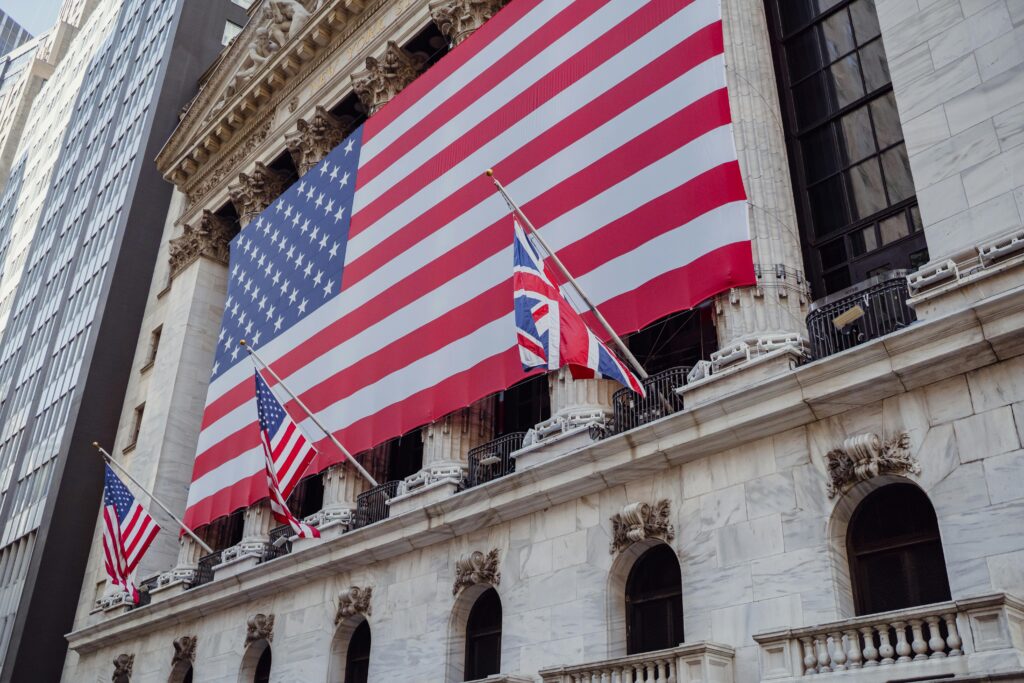 photograph of the New York Stock Exchange facade, featuring a massive American flag draped across the front and multiple Union Jack flags displayed on poles, with ornate architectural details and a modern skyscraper in the background.